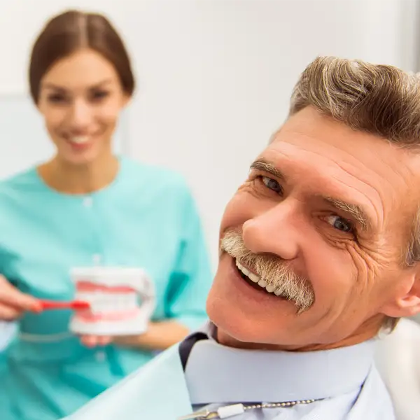 Older man smiling while dental hygienist demonstrates how to brush dentures