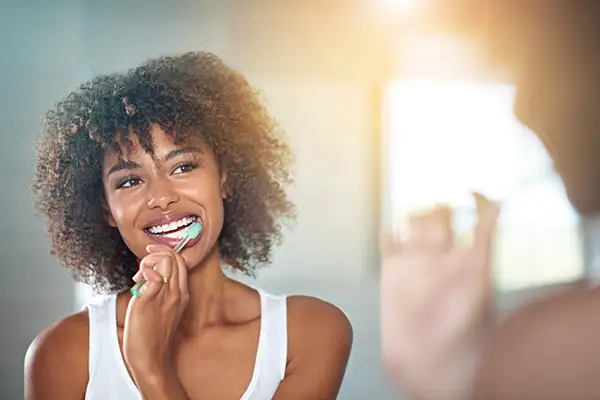 Smiling woman with curly hair brushing her teeth in a sunlit bathroom, emphasizing dental hygiene and oral care.